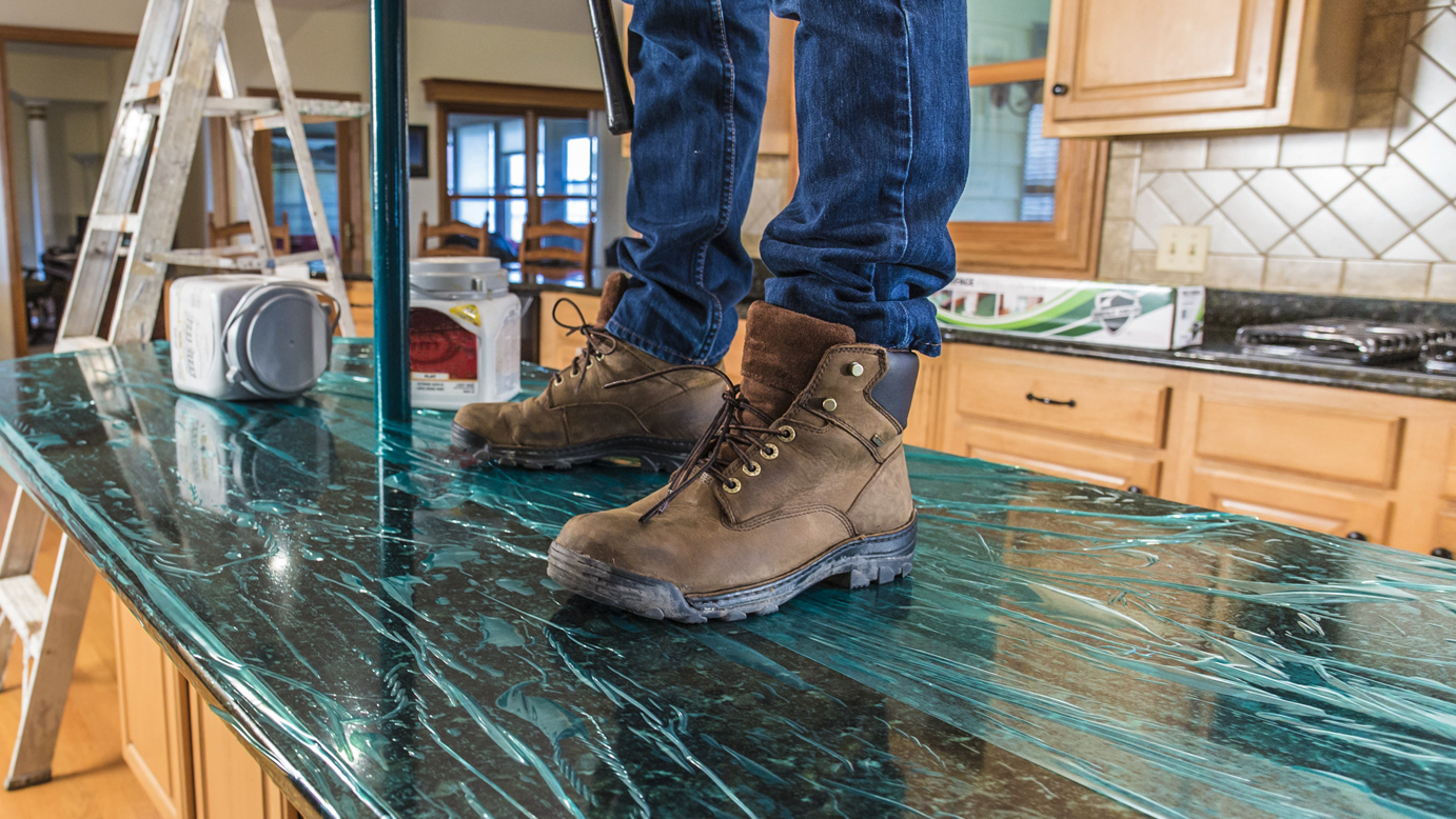 a close up of a person with work boots standing on a green, self adhesive surface protection film on a granite countertop.