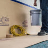person holding a 5 gallon bucket walking past equipment rest against builder board floor and wall protection
