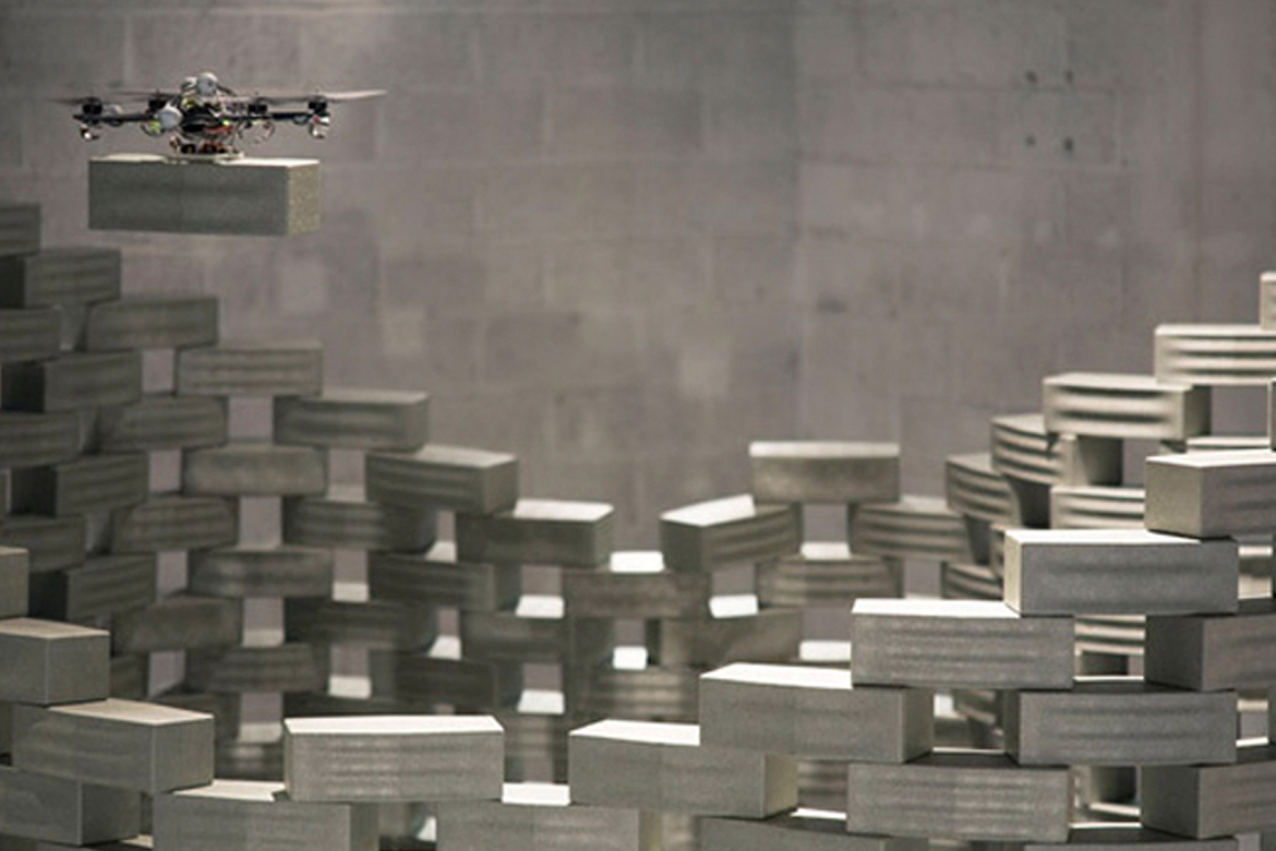 A drone holding a grey brick over a brick structure.