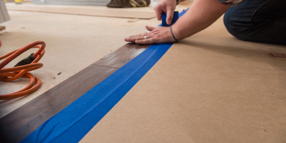 Close-up picture of a man while adhering a protective surface to the wooden floor with blue tape.